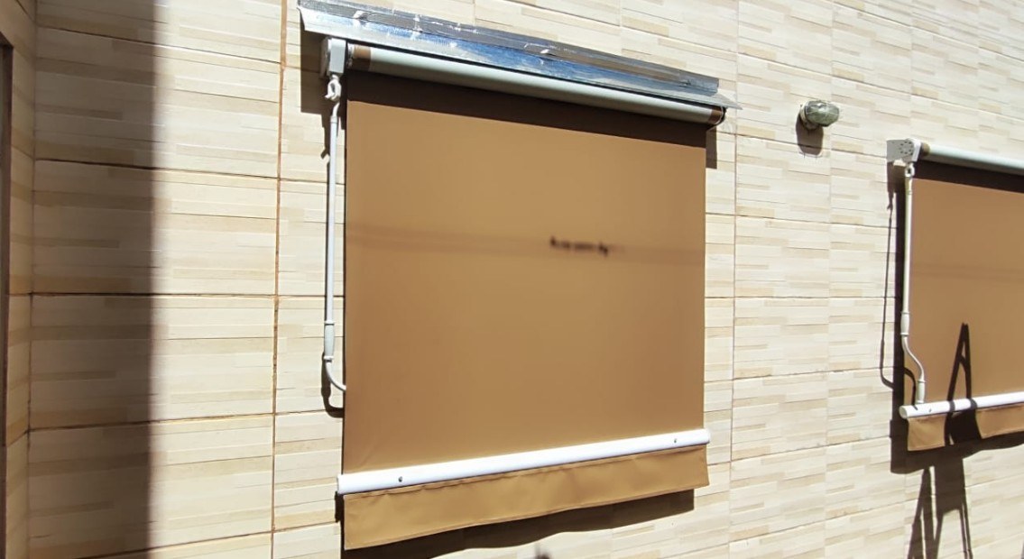 Rolô externo caramelo baixado do lado de fora de uma janela, numa parede de revestimento claro.