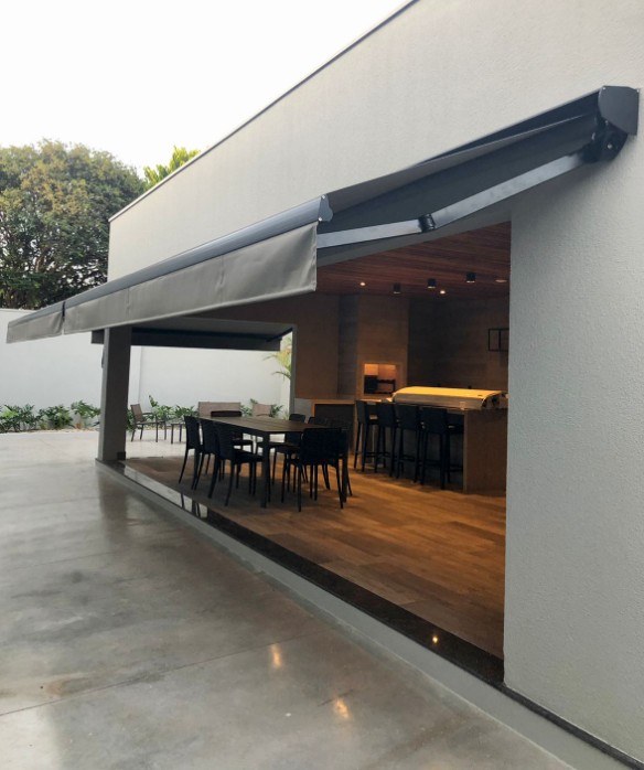Toldo articulado cinza aberto sobre a área gourmet de uma casa, com mesa e cadeiras pretas embaixo.