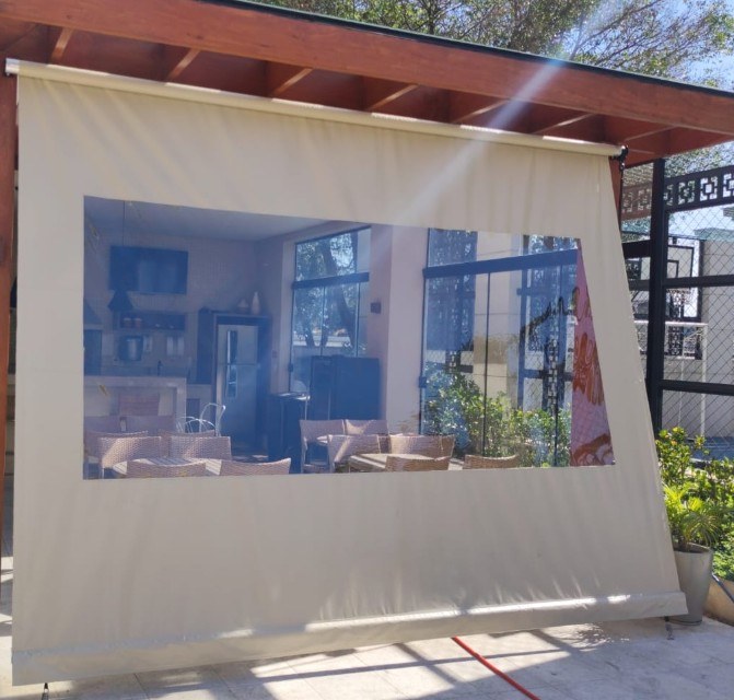 Toldo cortina creme baixado sob uma cobertura de madeira, com um grande visor transparente mostrando a área gourmet.